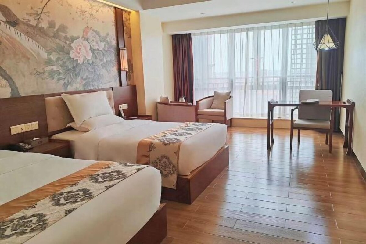 Ein Hotelzimmer mit zwei Betten, einem Schreibtisch, einem Sessel, einem großen Fenster mit Vorhängen und einem Wandbild.