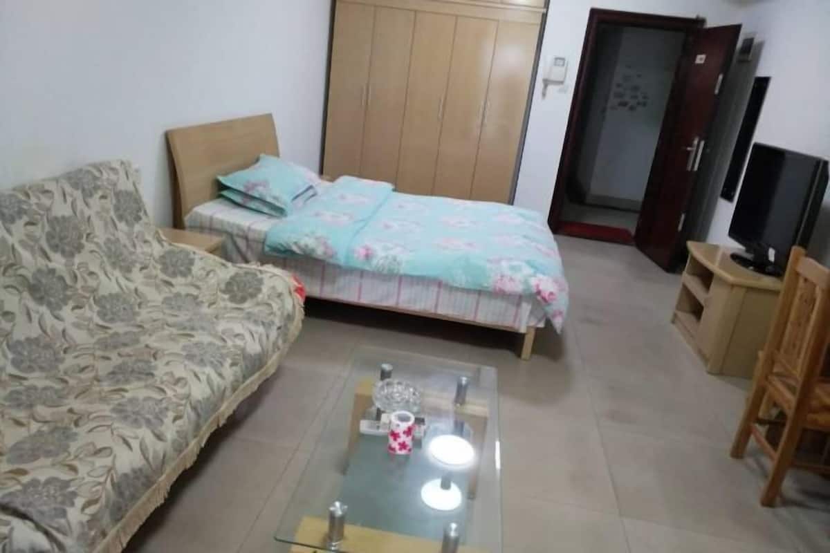 A room with a sofa, a bed with floral bedding, a glass coffee table with decorative items, a wardrobe, and a television.