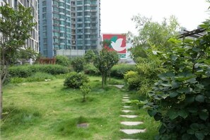 Un sentier bordé d’herbe, avec de petits buissons, et un grand immeuble en arrière-plan.