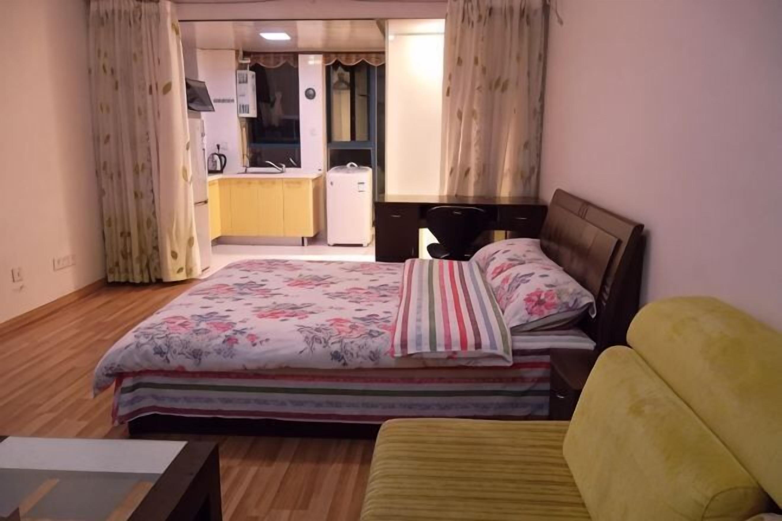 Une chambre à coucher avec un lit, un fauteuil jaune, un plancher en bois, un bureau et, au fond, une petite cuisine.