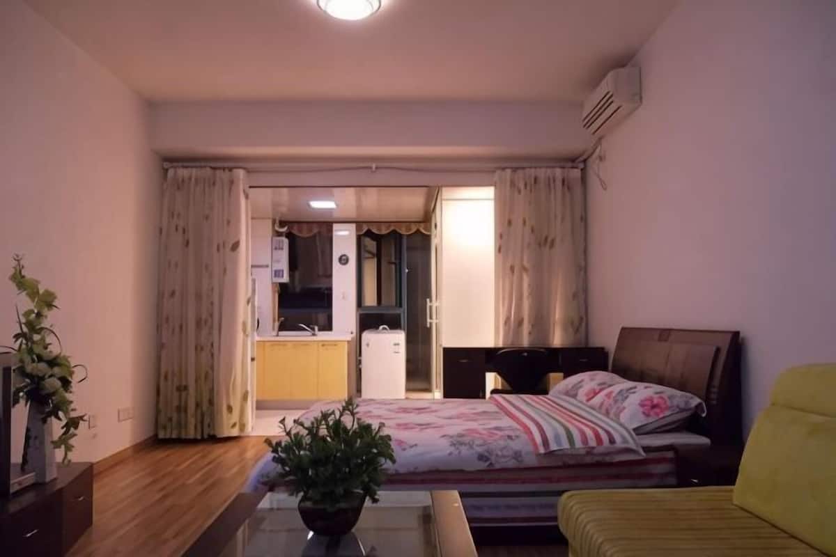 A bedroom with a bed, a bedside table, a chair, a potted plant, and a view of a kitchen and bathroom through an open door.