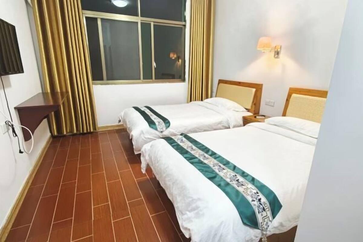 Habitación de hotel con dos camas, una ventana con cortinas, un televisor de pantalla plana y una lámpara fijada a la pared.