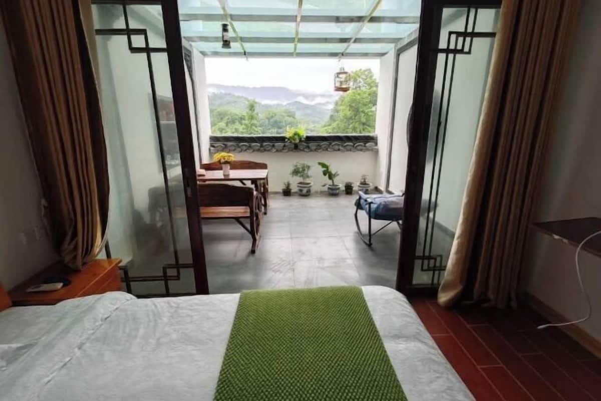 Una habitación con cama, una mesa con sillas, un balcón con plantas en macetas y vistas a zonas verdes.