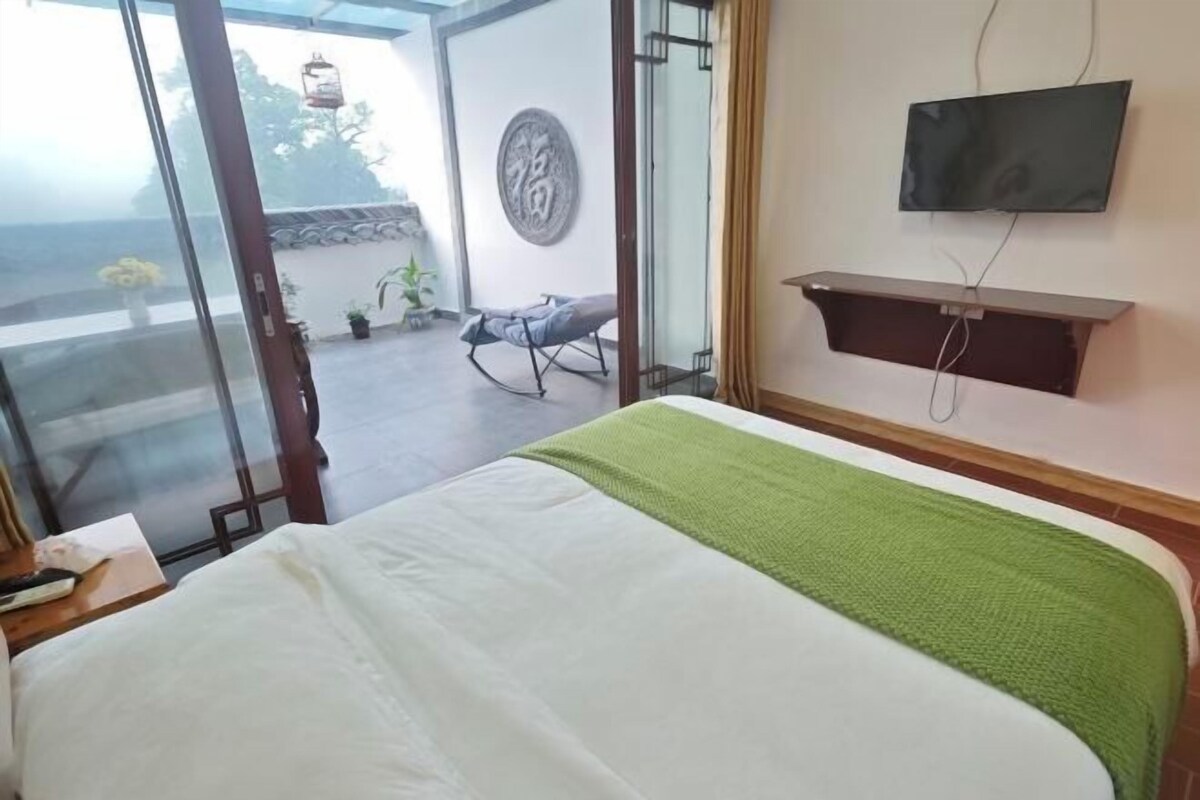 Quarto de hotel com cama, televisão fixada na parede, porta de vidro deslizante para área externa e uma pequena mesa com cadeira.