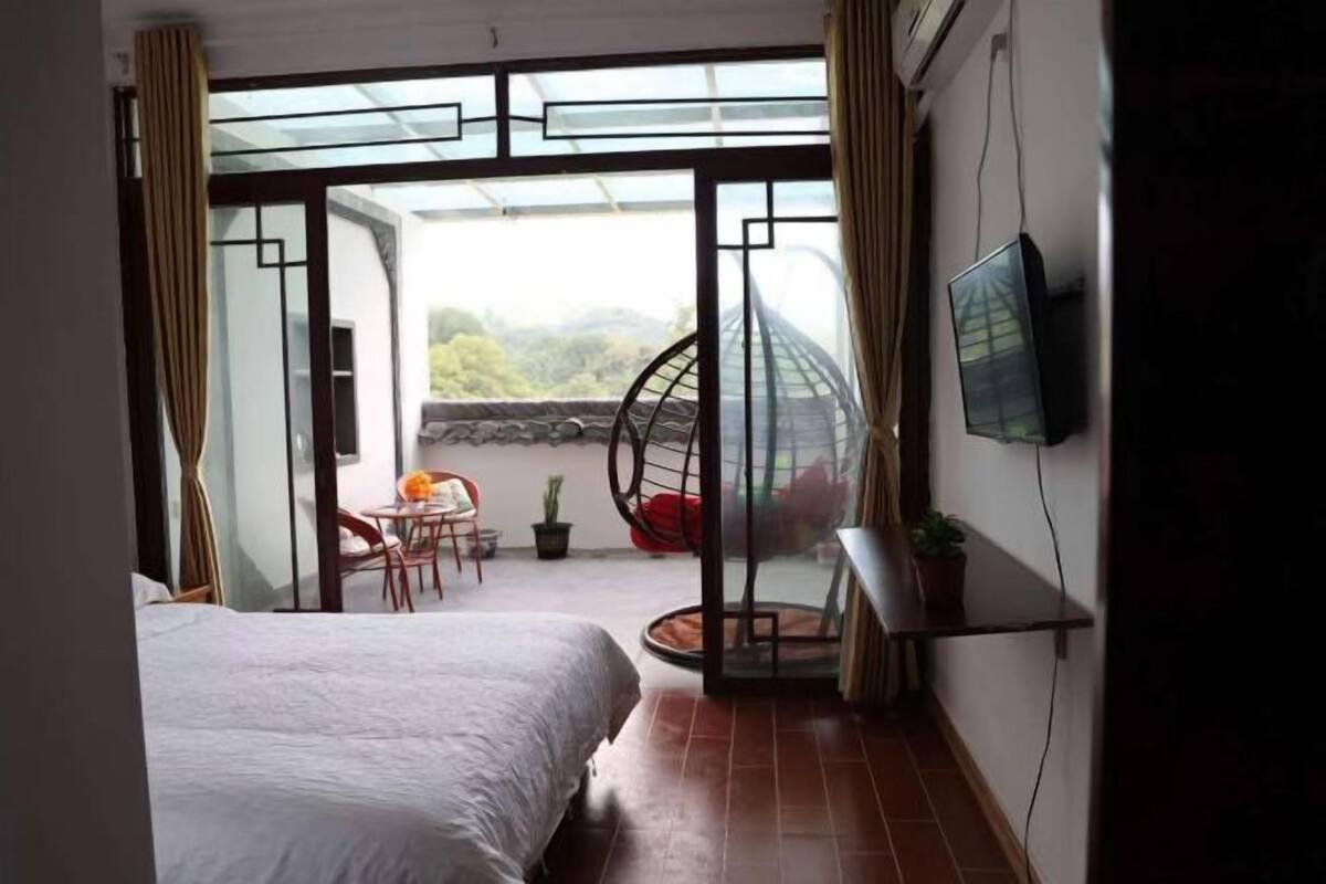 Un dormitorio con cama, un balcón con silla, televisión y vista a la vegetación.