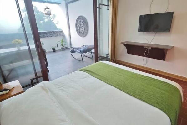 Quarto de hotel com uma cama, televisão fixada na parede e varanda com cadeira e plantas.