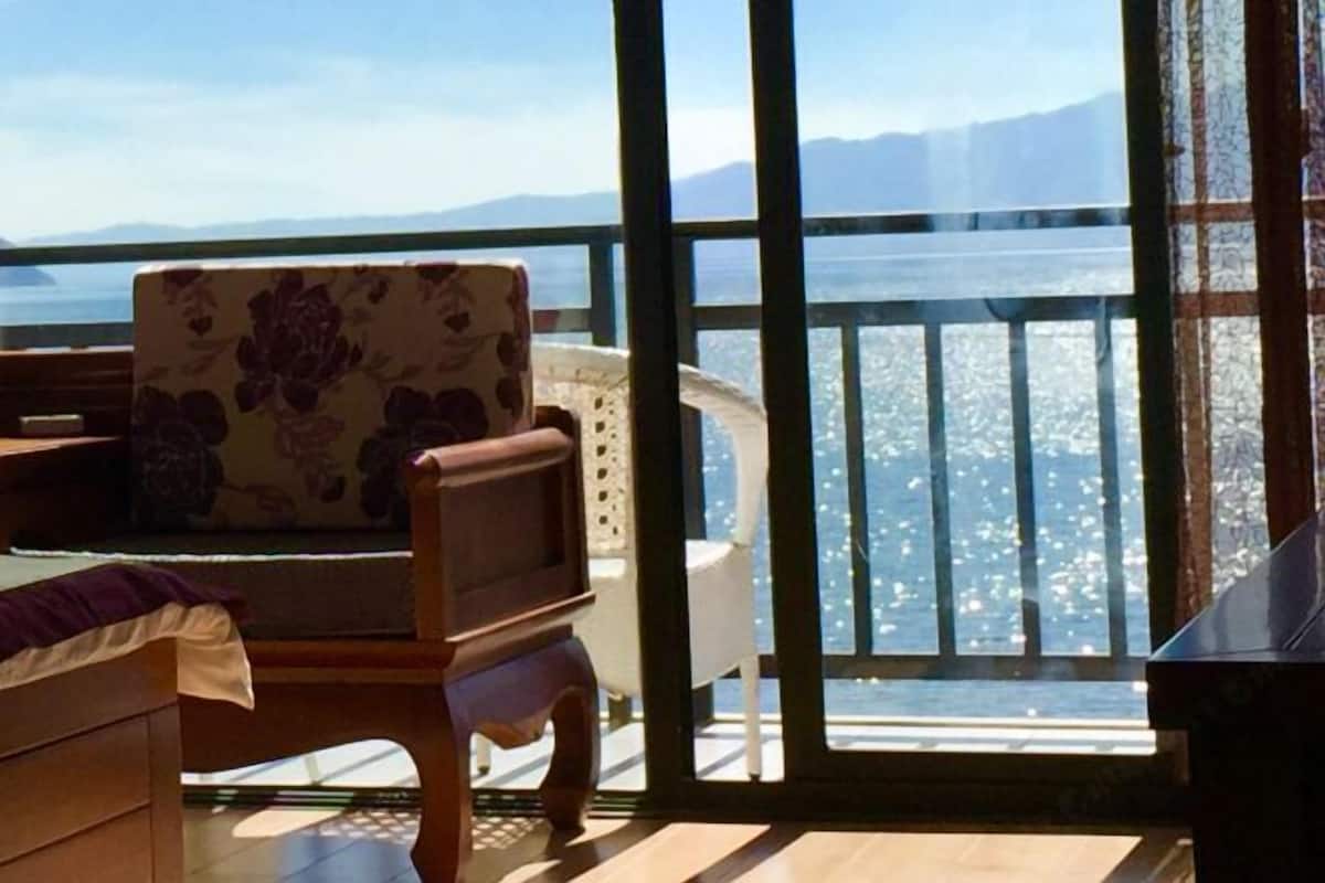 A balcony with a wooden chair, a white sofa, and a view of a body of water and mountains.