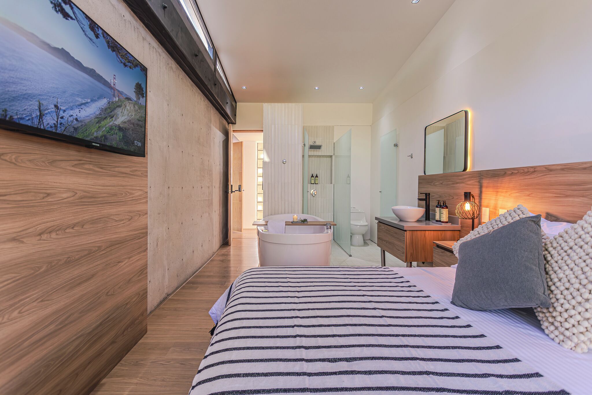 A modern bedroom with a large bed, wooden flooring, a TV on the wall, a bathroom with a glass shower, and a wooden vanity.