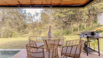 Eine Terrassensitzgelegenheit im Freien mit Korbstühlen, einem Glastisch, einem Grill und Blick auf eine natürliche Landschaft.
