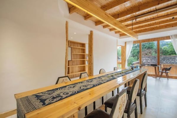 Una larga mesa de madera con sillas, un mantel decorado y vistas a un jardín a través de amplios ventanales.