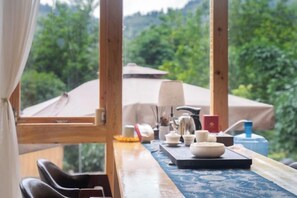 A dining area with a table set for two, a window with curtains, and a view of greenery outside.