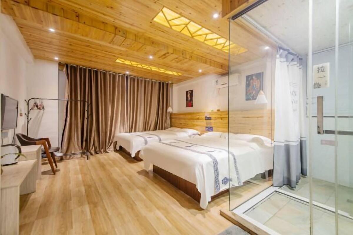 Hotellihuone, jossa on puulattia, kolme sänkyä, työpöytä ja lasiseinä.