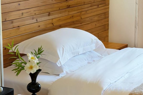 Una cama con sábanas blancas, cabecera de madera y un florero sobre la mesita de noche.