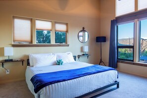 Une chambre à coucher avec un lit, une fenêtre avec des stores, une table de chevet avec une lampe et un miroir fixé au mur.