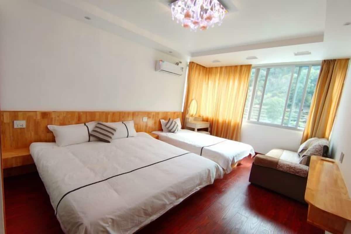 Habitación de hotel con dos camas, cabecero y banco de madera, una mesita pequeña y un ventanal grande con cortinas.
