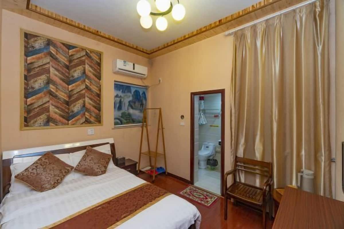 Una habitación de hotel con cama, una silla de madera, una mesa, un cuadro y una ventana con cortinas.
