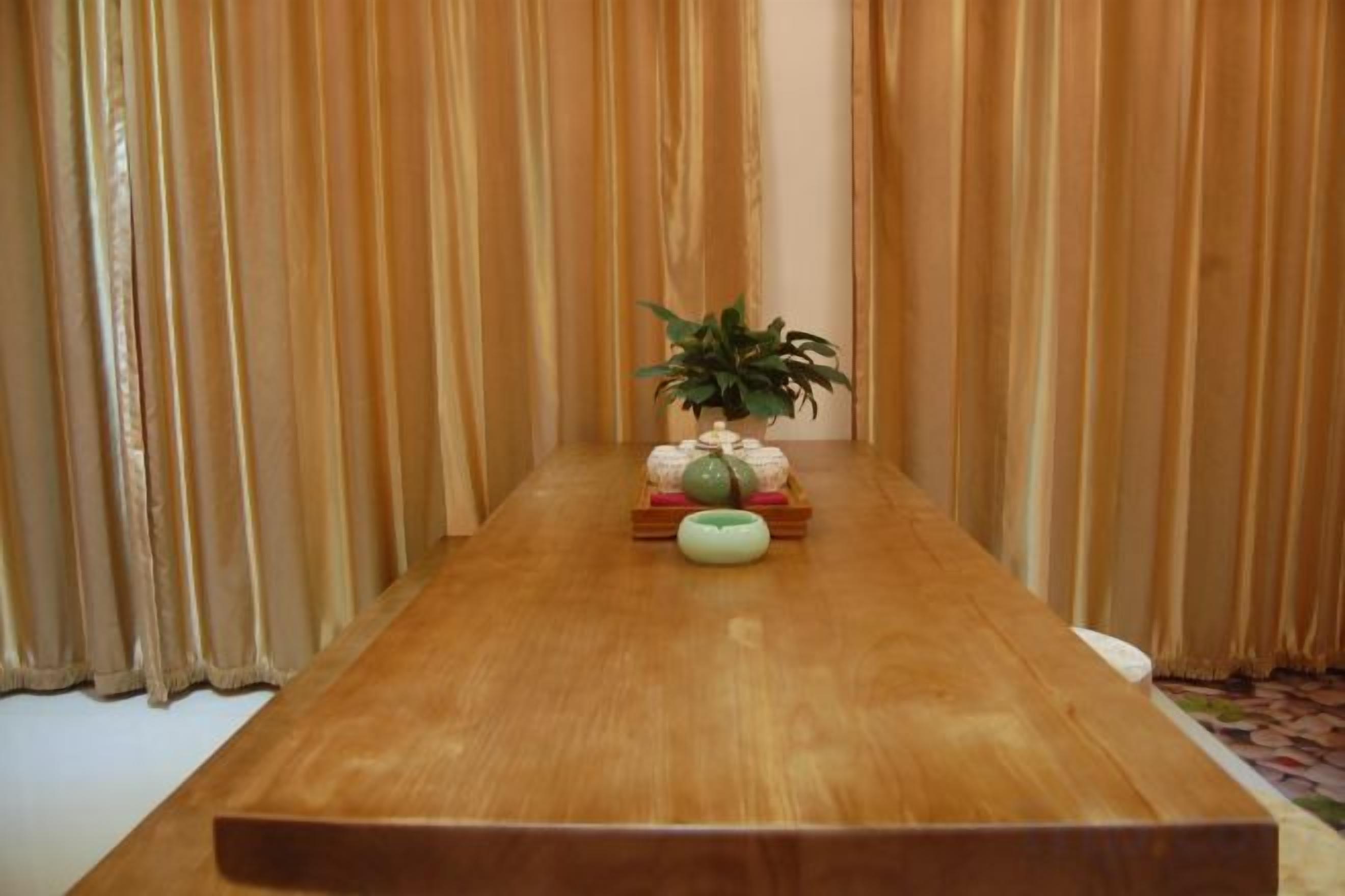 Una mesa de madera con una planta en maceta y un bol en el centro, frente a un fondo de cortinas verticales.