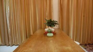 Una mesa de madera con una planta en maceta y un bol en el centro, frente a un fondo de cortinas verticales.