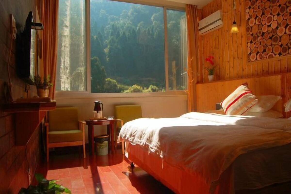 Una habitación con cama, una silla, una mesita, un ventanal con vista a árboles y una pared de madera con diseño de troncos.