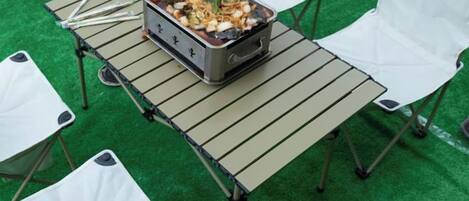 Una mesa con un recipiente de comida y palillos, rodeada de sillas blancas sobre una alfombra verde.