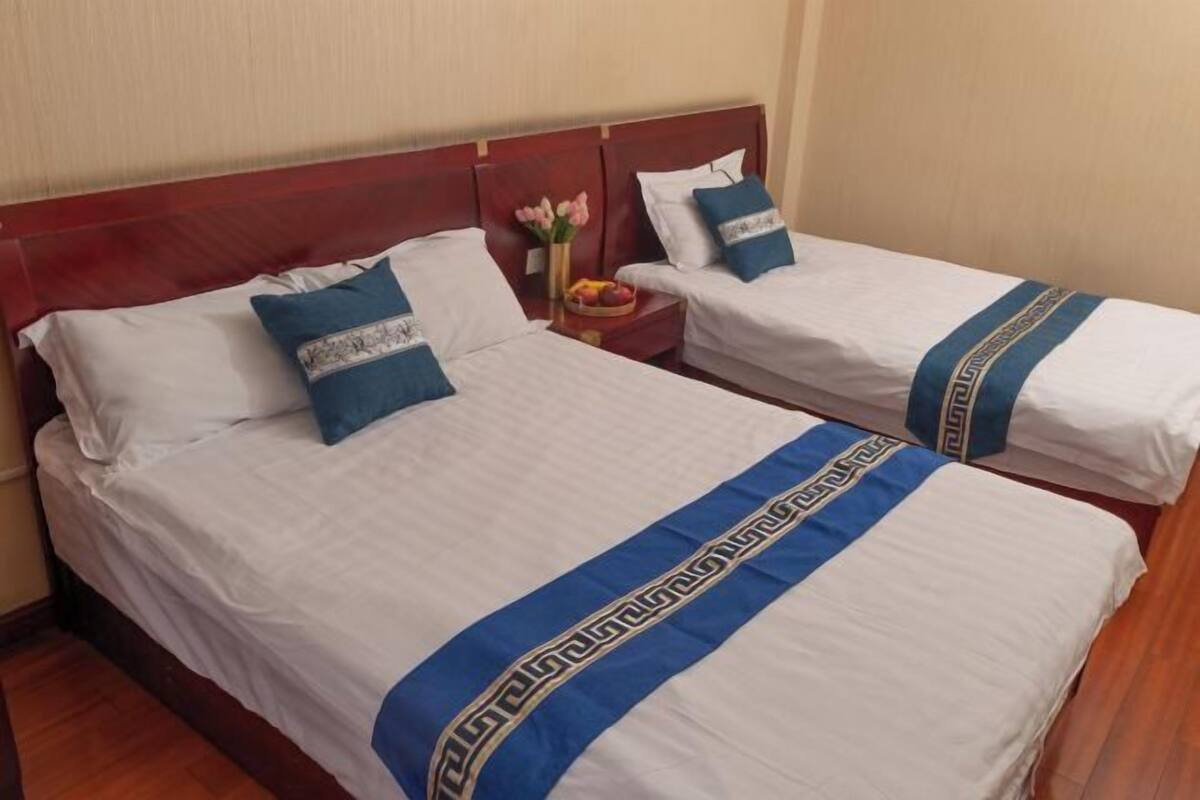 A hotel room with two single beds, each with a white bedspread and a blue decorative stripe. There is a wooden headboard, a small bedside table with a vase of flowers, and a fruit bowl.