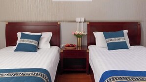 Une chambre d’hôtel avec deux lits, chacun doté d’une tête de lit en bois et de linge de lit blanc. On y trouve une petite table de chevet avec un vase de fleurs et un panier de fruits. Un tableau encadré représentant une jetée au-dessus de l’eau est accroché au mur, au-dessus des lits.