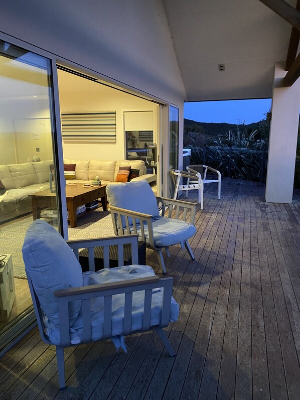 A covered outdoor seating area with white cushioned chairs and a wooden table, overlooking a garden with tall plants and a view of the hills.