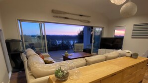 Un soggiorno moderno con un grande tavolo da pranzo in legno, un divano beige e una porta scorrevole in vetro che dà su un balcone con vista sul tramonto.
