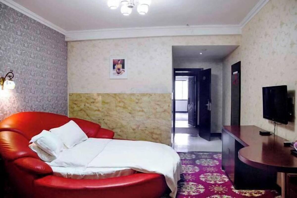 Una habitación de hotel con una silla de cuero rojo, una cama con ropa blanca, un televisor y una alfombra estampada.