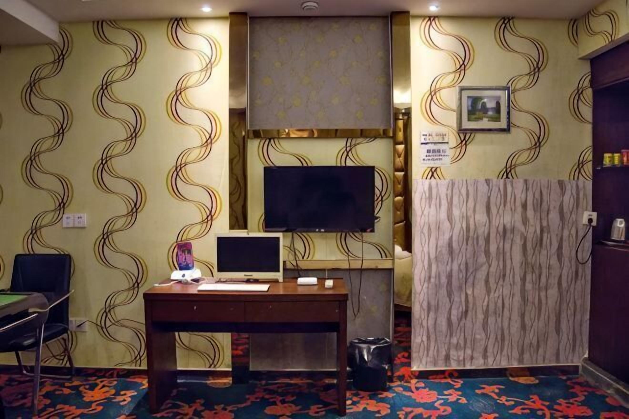 A hotel room with a desk, two monitors, a television, a patterned wall, and a carpet.