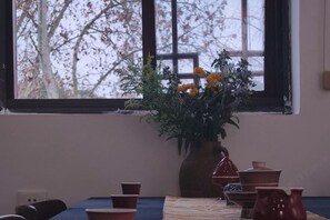 Un comedor con una mesa preparada para dos personas, un florero con flores y un ventanal con vista a árboles.