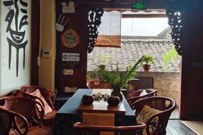 An indoor seating area with wicker chairs and a table, a decorative wall with calligraphy, and a view of a traditional tiled roof through an open doorway.