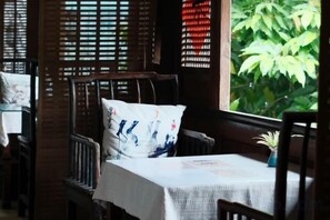 Un área de comedor con muebles de madera, una mesa con mantel blanco y vistas a la vegetación exterior.