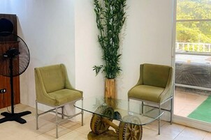 A room with two green chairs, a glass table, a tall bamboo plant, and a fan.