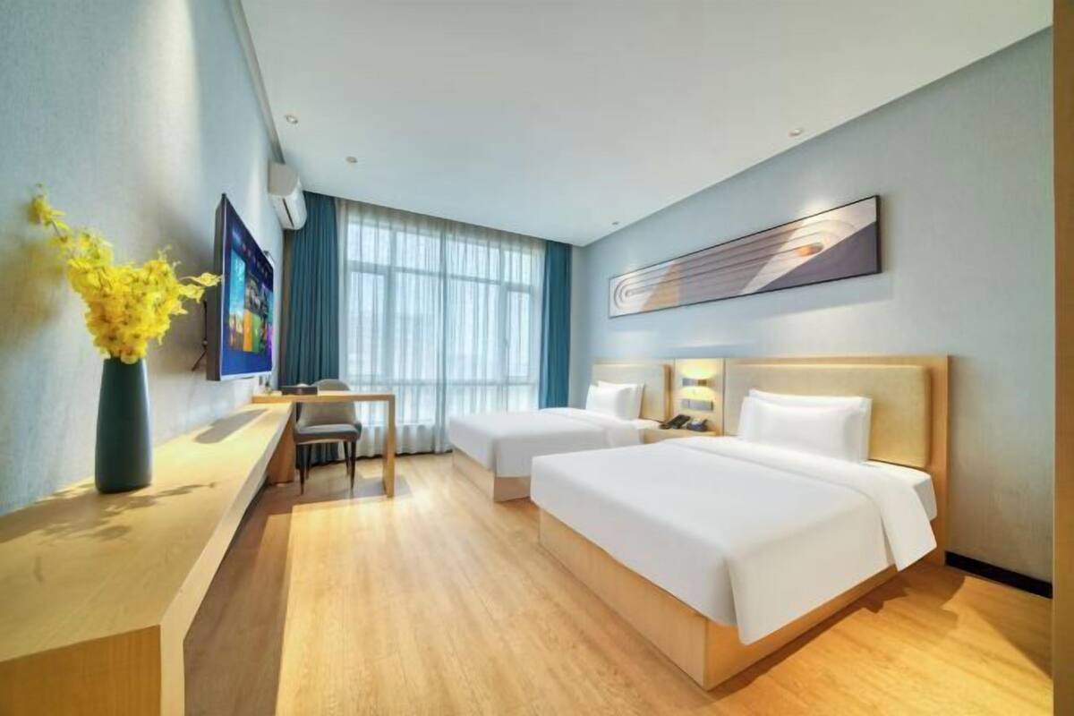 A hotel room with a desk, two beds, a large window, and a wall-mounted artwork.