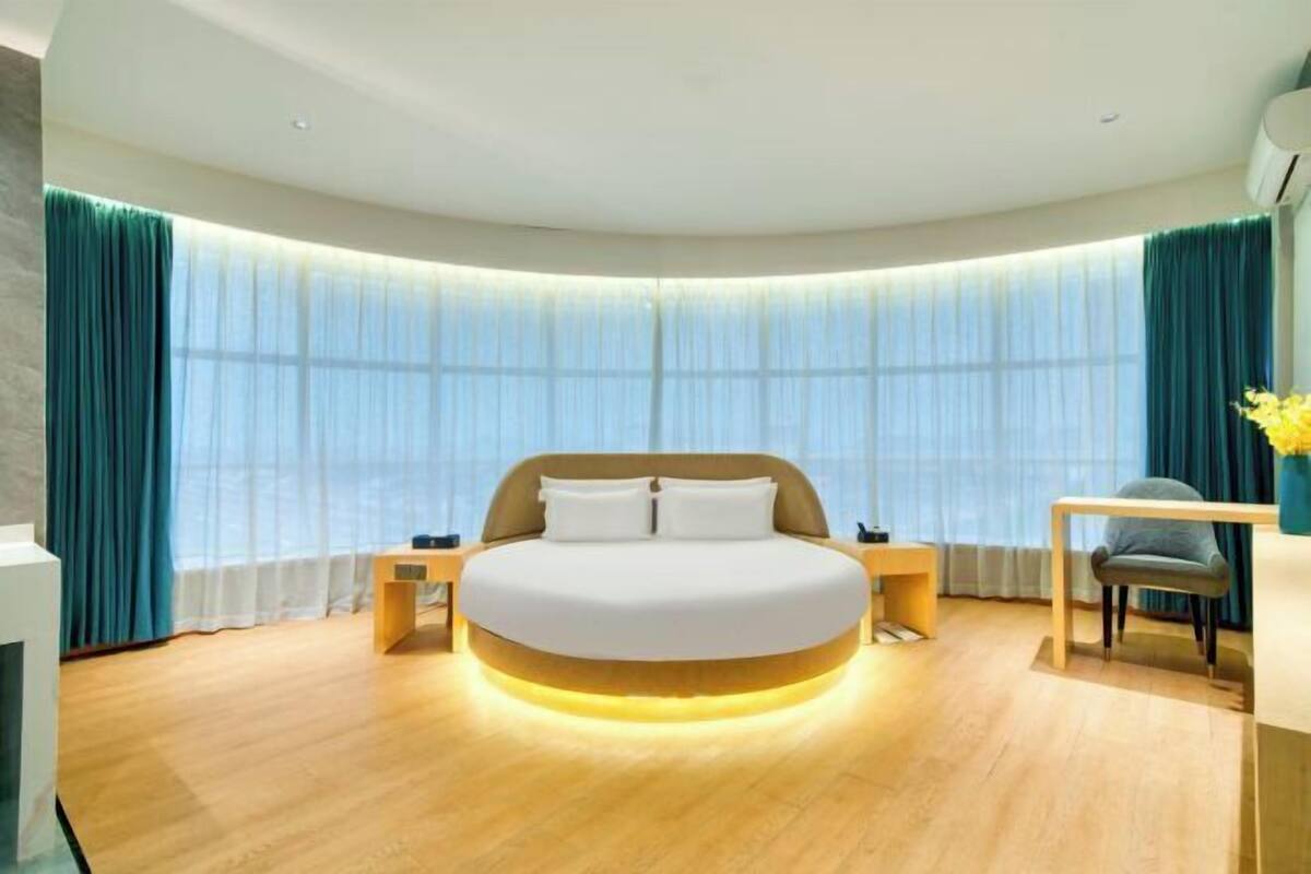 A modern hotel room with a curved bed, a desk, and a view of the city through large windows.