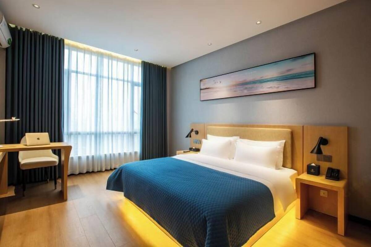 A hotel room with a bed, a desk, a chair, a nightstand, a lamp, and a painting on the wall.