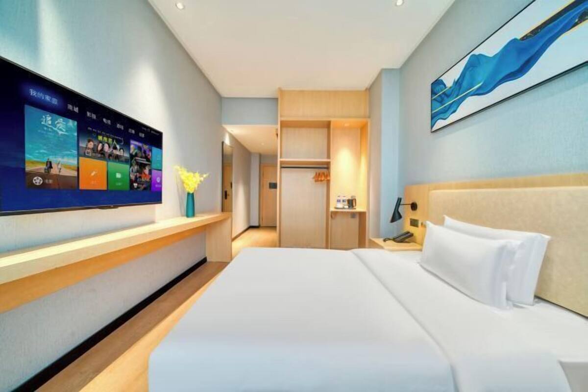 A hotel room with a large bed, a TV mounted on the wall, a desk with a lamp, and a wardrobe.