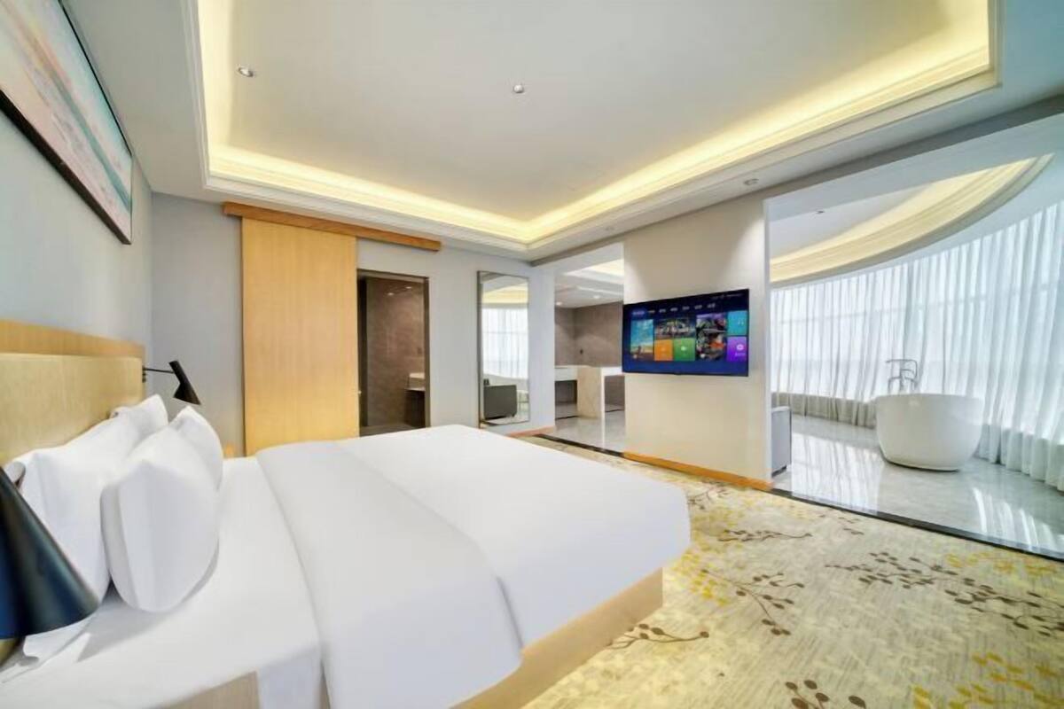 A hotel room with a bed, a TV, a large window with curtains, and a bathtub.