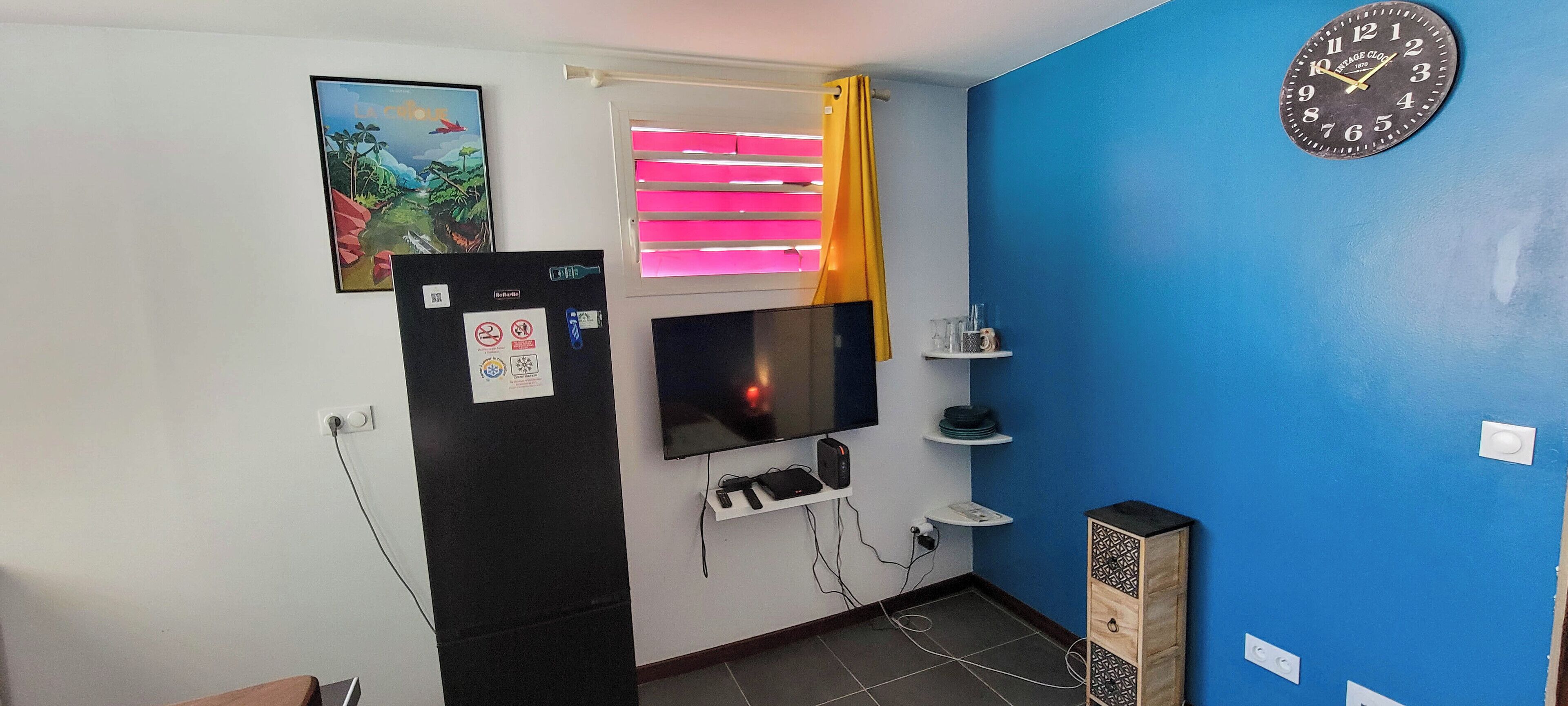 A room with a TV mounted on the wall, a small shelf with glasses, and a clock.