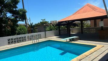A swimming pool with a tiled deck, a covered seating area, and a view of surrounding greenery.