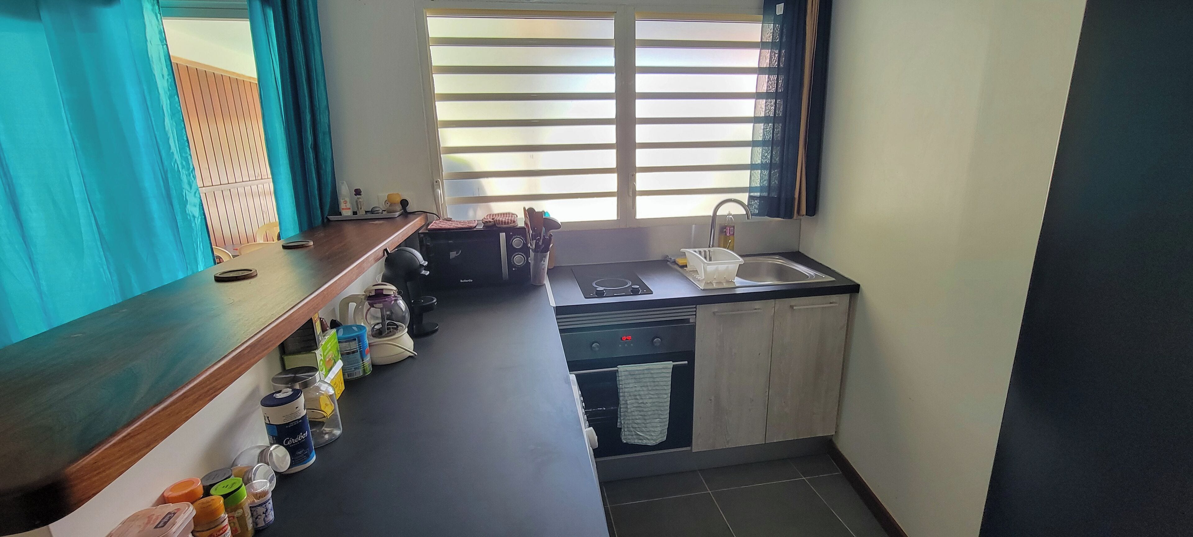 A compact kitchen with a sink, stove, and cabinets. There are various kitchen items on the counter, including a kettle, a coffee maker, and containers.