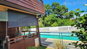 A wooden exterior wall with a sliding glass door, a small table with chairs, and a view of a swimming pool. - Sweet Dreams Bourda - Sérénité & Piscine (Cayenne)