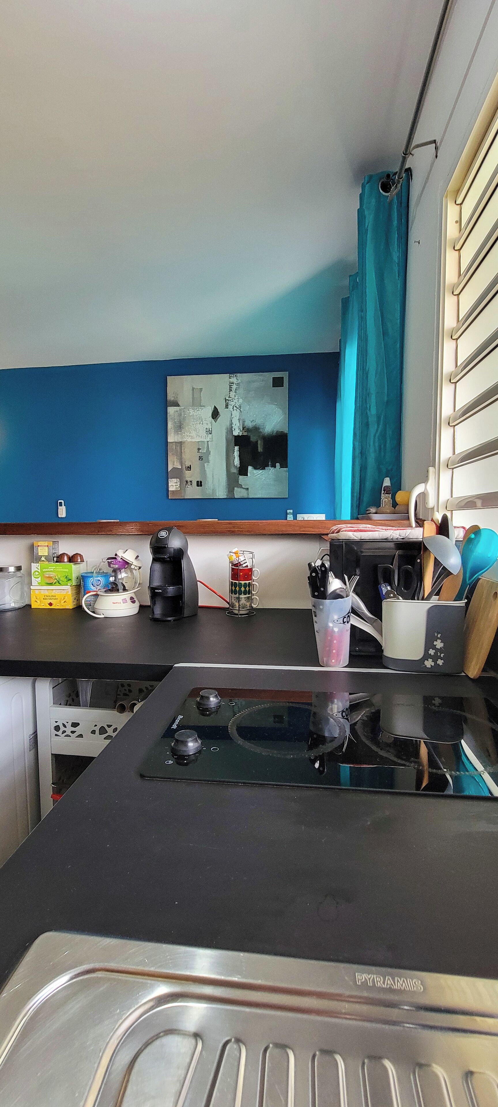 A kitchen with a black countertop, a coffee maker, a toaster, and a blender. There is a window with blue curtains and a painting on the wall.