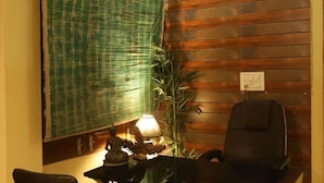 A room with a wooden wall, a green patterned curtain, a desk with a lamp, a chair, and a potted plant.