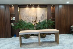 A reception area with a long wooden reception desk, two potted plants, a wall with a decorative panel, and a bookshelf. - Yue Cheng Commercial Hotel (Anping)