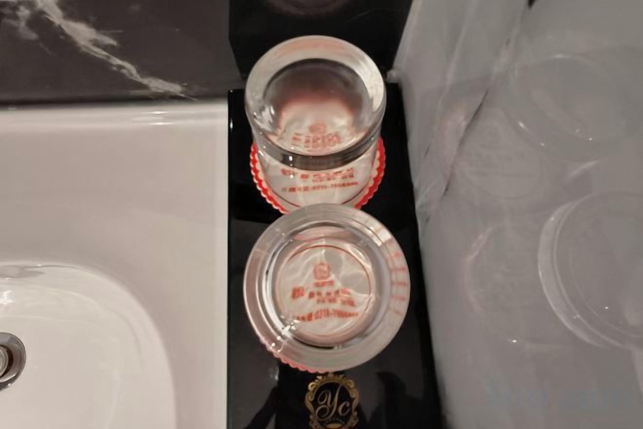 Dos botellas de vidrio transparente con etiquetas rojas sobre una superficie reflectante.