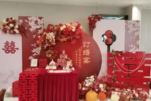 Un espacio decorado para eventos, con arreglos florales, una mesa con objetos y un telón de fondo rojo con caracteres chinos.