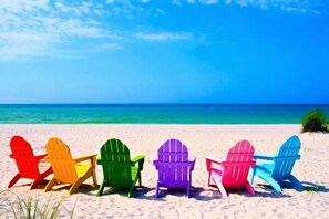 Une rangée de chaises Adirondack aux couleurs vives sur une plage de sable, avec un ciel bleu clair et l’océan en arrière-plan.