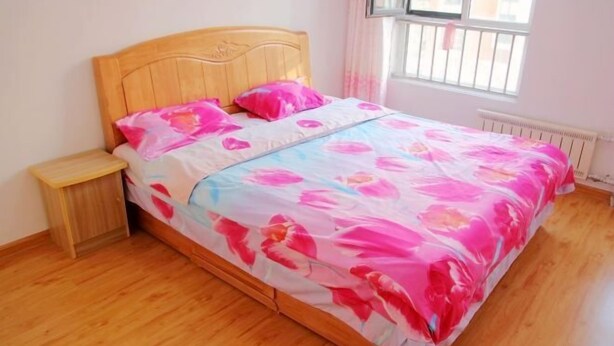 A single bed with a floral bedspread, a wooden headboard, a nightstand, and a window with a view of buildings.